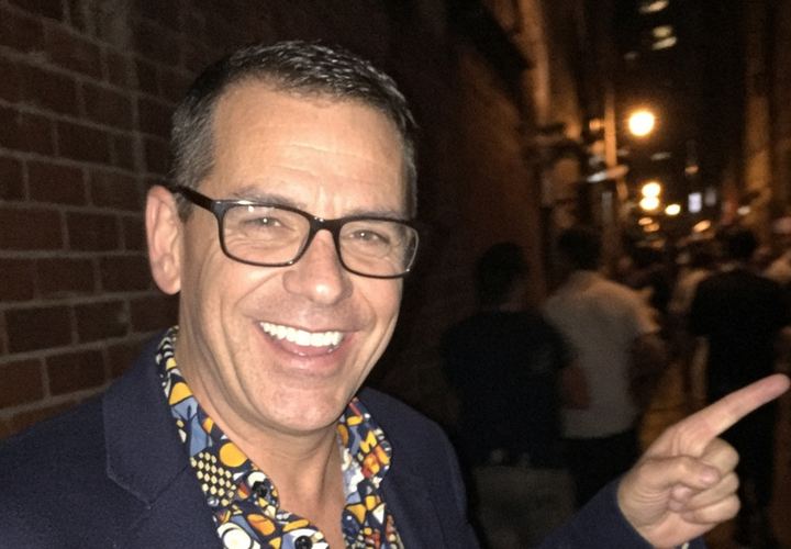 Greg, grinning in glasses, a navy blazer and a loud patterned shirt, pointing down a busy night-time laneway at the place he swears he knows
